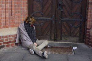 Eine zusammengesunkene Person, in schäbiger Kleidung, deren Gesicht man nicht erkennt, vor einer Kirchentür