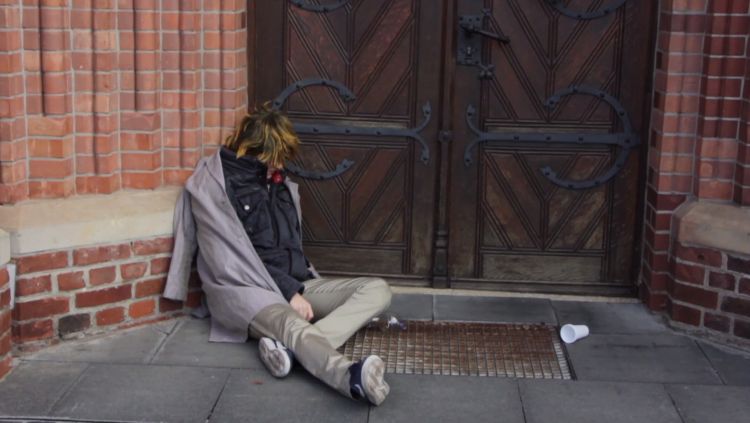 Eine zusammengesunkene Person, in schäbiger Kleidung, deren Gesicht man nicht erkennt, vor einer Kirchentür