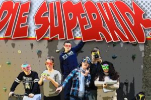 Jugendliche vor einer Kletterwand, mit Superheldenmasken im Gesicht. Stark comicartig verfremdet. Typische Comic-Schrift: "Die Superkids"