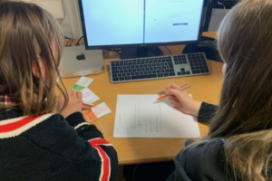 Zwei Personen sitzen an einem PC und schreiben etwas auf ein Blatt Papier