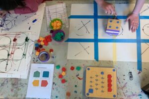 Auf einem Tisch liegen große Blätter, auf die Raster geklebt sind und die von den Makey-Makeys bemalt werden. Kinderhände arbeiten daran. Außerdem verstreut viele bunte Steine der Makey-Makeys.