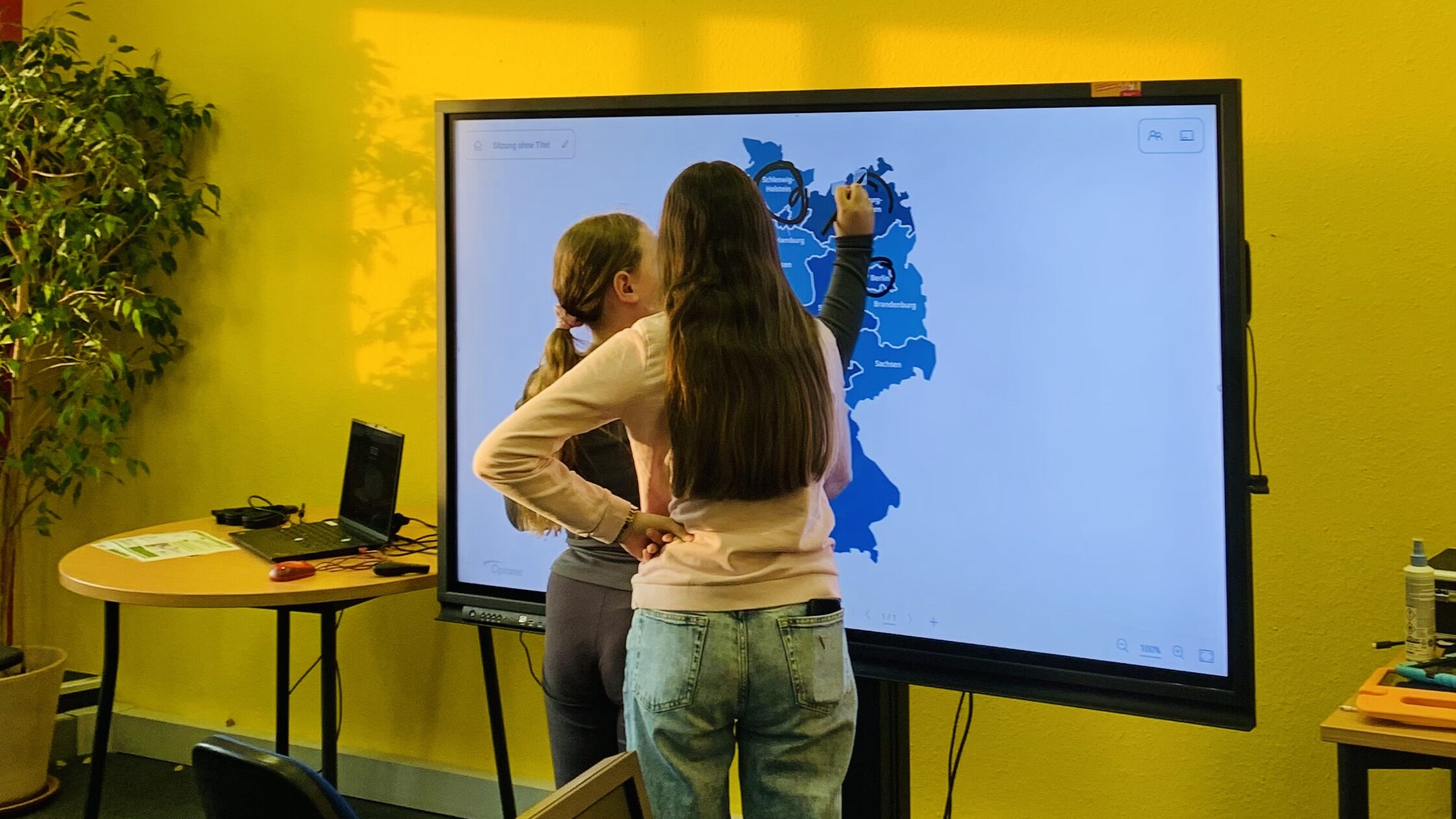 Zwei Mädchen stehen vor einem großen Bildschirm. Auf dem Bildschirm sieht man eine Landkarte von Deutschland. Das eine Mädchen hat einen Stift in der Hand und kreist damit digital Orte auf der Landkarte ein.