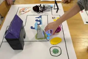 Hand bewegt einen kleinen blauen Ozo-Bot auf einer selbst gemalten Karte mit Hindernissen und 3D-Elementen auf einem Tisch.