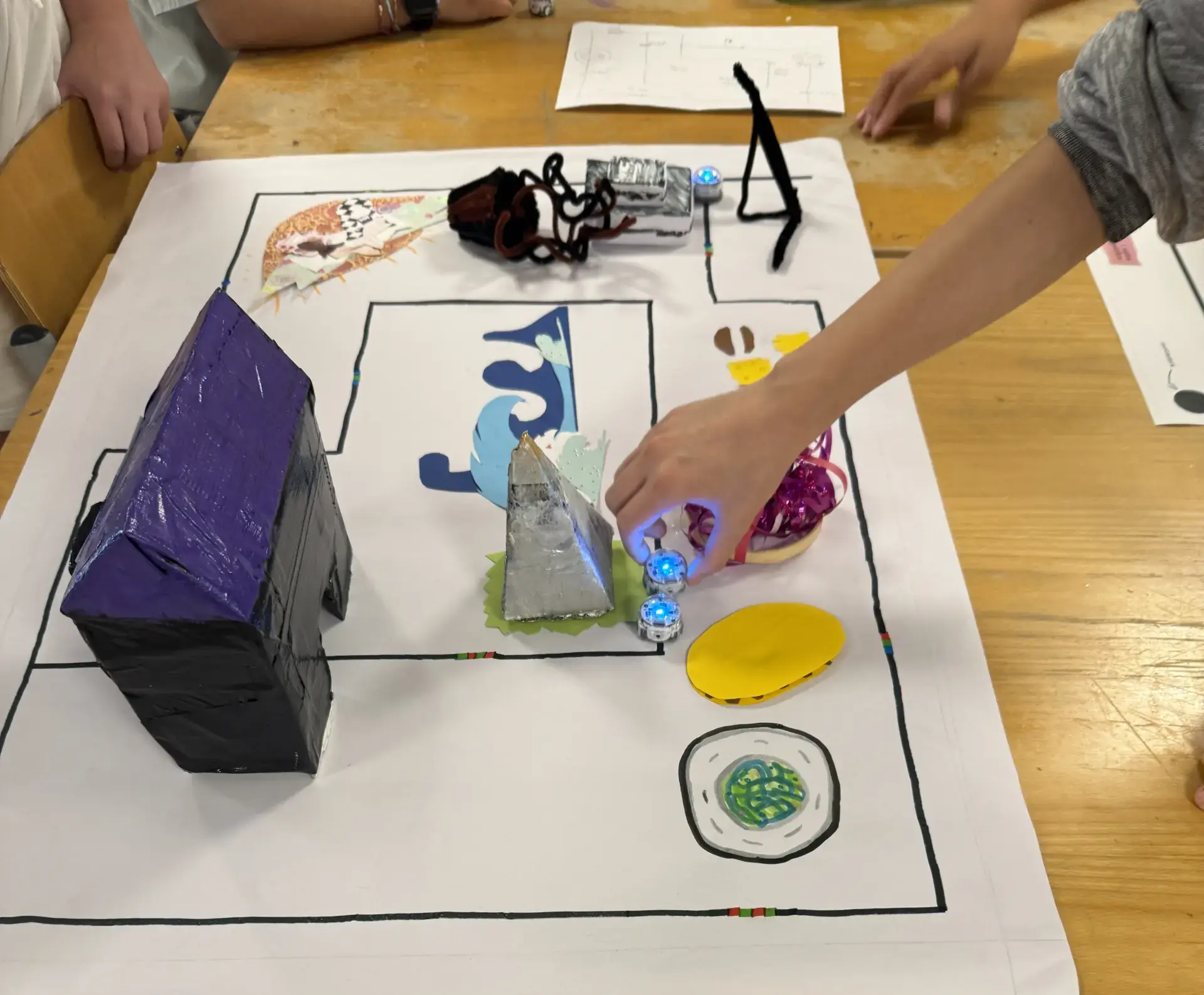 Hand bewegt einen kleinen blauen Ozo-Bot auf einer selbst gemalten Karte mit Hindernissen und 3D-Elementen auf einem Tisch.