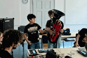 Zwei Jungen stehen in einem Raum, einer hält eine schwarze Tube, der andere Junge hält ein Mikrofon. Mehrere Personen sitzen an einem Tisch mit Laptops und Musikequipment.