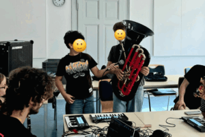 Zwei Jungen stehen in einem Raum, einer hält eine schwarze Tube, der andere Junge hält ein Mikrofon. Mehrere Personen sitzen an einem Tisch mit Laptops und Musikequipment.