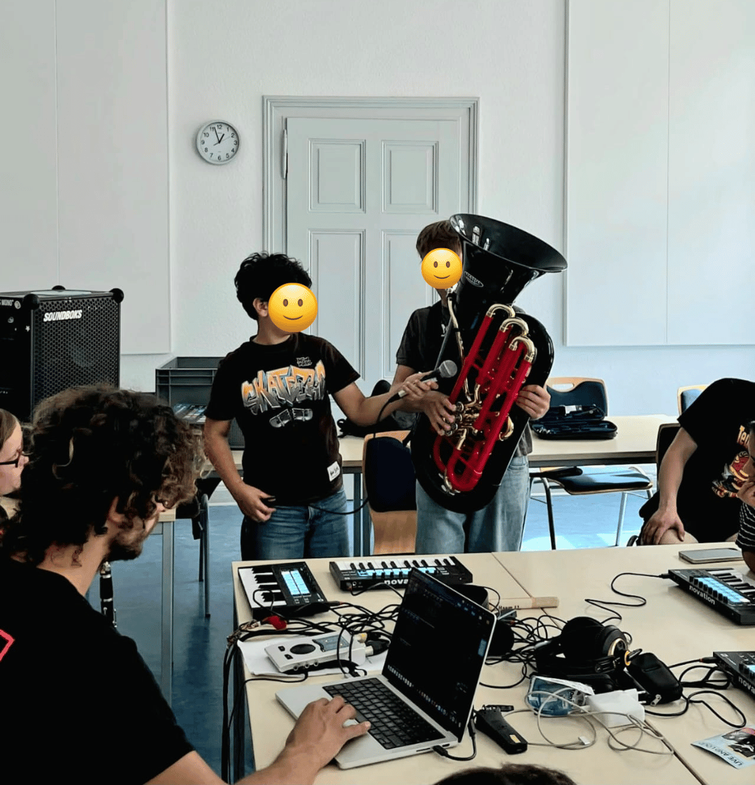 Zwei Jungen stehen in einem Raum, einer hält eine schwarze Tube, der andere Junge hält ein Mikrofon. Mehrere Personen sitzen an einem Tisch mit Laptops und Musikequipment.
