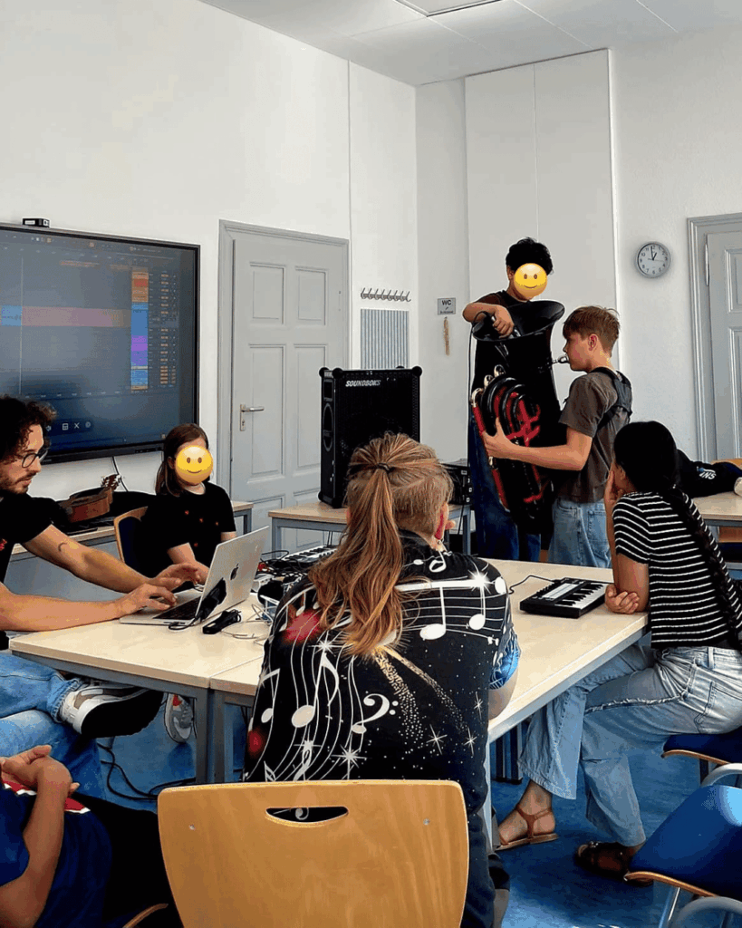 Fünf Jugendliche in einem Soundstudio, zwei sitzen am Tisch mit Laptop und Keyboard, zwei stehen und halten ein großes Blasinstrument.