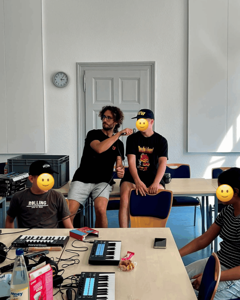 Zwei Jugendliche sitzen an einem Tisch mit zwei Keyboards, ein Workshopleiter hält einem anderen Jungen ein Mikrofon hin.